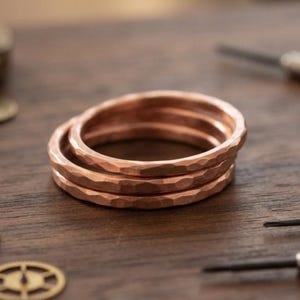 Puede incluir: Un conjunto de tres anillos metálicos finos de color cobre con una superficie martillada y texturizada. Los anillos son circulares, de ancho constante, con un acabado metálico cálido y un patrón facetado en la banda exterior.