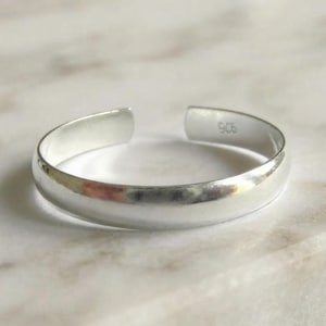 Könnte beinhalten: Ein polierter, silberfarbener offener Ring mit einer glatten, abgerundeten Oberfläche. Auf der Innenseite des Rings befindet sich die Prägung 925, die auf die Metallzusammensetzung hinweist. Der Ring hat eine Öffnung zur Größenanpassung.