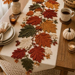 Può includere: Un runner da tavola beige con una composizione decorativa centrale di foglie autunnali all'uncinetto nei toni del ruggine, verde oliva, crema e marrone, arricchita da piccole ghiande all'uncinetto lungo una struttura a tralcio.