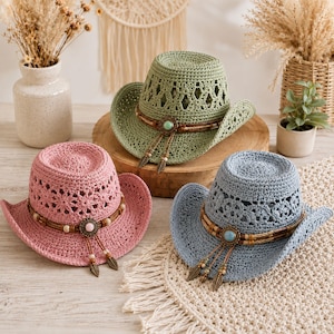 May include: Three crocheted cowboy hats in sage green, dusty pink, and slate blue. Each hat features an open-weave crown, a curved brim, and a decorative band made of brown cord, wooden beads, a central circular metal concho, and dangling leaf charms.
