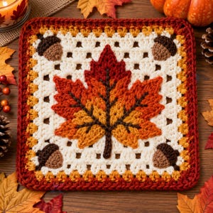 May include: A square crochet granny square featuring a large central maple leaf in shades of red, orange, and yellow. The background is off-white with four brown acorns placed in each corner. The border is a deep red with a yellow trim.