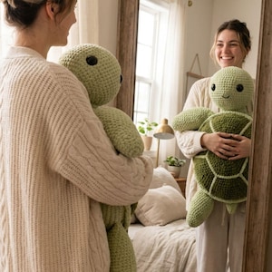 Puede incluir: Un peluche de tortuga tejido a ganchillo a mano, con cabeza y extremidades de color verde claro y un caparazón segmentado de color verde oscuro. La tortuga tiene dos ojos redondos de plástico negro y una sonrisa sencilla bordada en la cara.