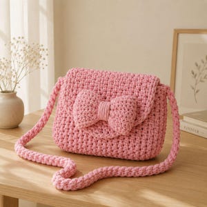 Può includere: Una borsa all'uncinetto rosa chiaro caratterizzata da un motivo a maglia spesso e testurizzato, con un fiocco all'uncinetto abbinato ben visibile sulla patta anteriore. La borsa include una lunga tracolla intrecciata ed è realizzata in filato morbido.
