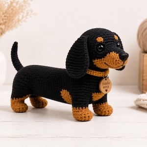 Puede incluir: Un juguete de teckel tejido a ganchillo con hilo negro y marrón, que presenta un collar amarillo y una etiqueta redonda de madera colgada del collar, grabada con el texto "made with" y un pequeño símbolo de corazón.