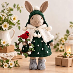 Peut inclure: Une poupée lapin tricotée aux longues oreilles, portant une cape à capuche vert foncé avec des pompons blancs, un châle blanc et une robe vert foncé à pois blancs. Le lapin tient une petite couronne marron avec des feuilles vertes et un oiseau rouge.