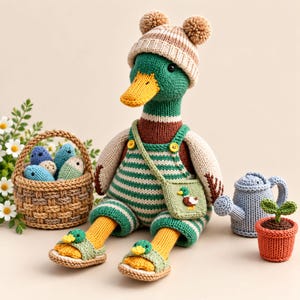 May include: A crocheted mallard duck doll wearing a striped beanie with two pom-poms, green and white striped overalls, and sandals with duck accents. It carries a small matching bag and sits beside a woven basket of fish and a blue watering can.