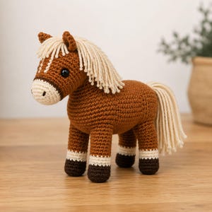 May include: A brown crocheted horse toy with a cream-colored muzzle, mane, and tail. The horse features dark brown hooves, cream-colored leg markings, and black safety eyes. The body is crafted from yarn with a visible stitch pattern throughout.