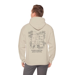 May include: A beige hooded sweatshirt featuring a black line art graphic on the back. The design displays a floor plan layout surrounded by various potted plants, with the text "Precision in engineering, patience in gardening." printed below.