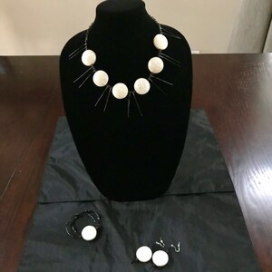May include: A black necklace with white circular accents and black linear extensions is displayed on a black mannequin. A matching bracelet and earrings with white circular accents are also visible on a black surface.