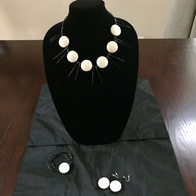 Puede incluir: Un collar negro con detalles circulares blancos y extensiones lineales negras se exhibe en un maniqu&iacute; negro. Tambi&eacute;n se ven una pulsera y pendientes a juego con detalles circulares blancos.