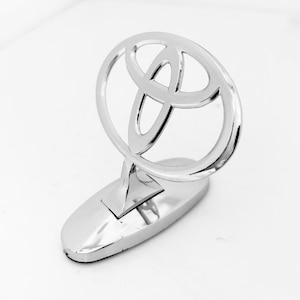 Könnte beinhalten: Ein poliertes Toyota-Emblem aus Chrom für die Motorhaube mit dem ikonischen Drei-Oval-Logo-Design. Das metallische Abzeichen ist auf einer glatten, ovalen Basis montiert und weist eine reflektierende, spiegelartige Oberfläche am gesamten Autozubehör auf.