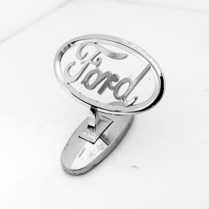 May include: A polished chrome Ford emblem bonnet mascot featuring the iconic script logo enclosed within an oval frame. The emblem is mounted on a matching chrome base, showcasing a reflective metallic finish and a classic automotive design.
