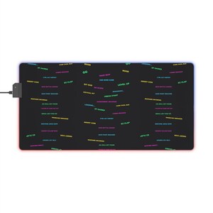 May include: A rectangular black gaming mouse pad featuring a repeating pattern of colourful neon gaming phrases including "GG," "NOOB," "LEVEL UP," "PRESS START," "LOADING," "XP GAINED," "INSERT COIN," "EZ CLAP," and "RESPAWN INCOMING" with an integrated power cord.