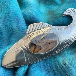 May include: A silver fish-shaped pendant with a textured surface and a clear, oval-shaped stone in the center. The fish has detailed fins and a tail, and is set against a blue and teal fabric background.