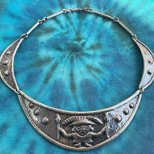 May include: A silver-toned necklace with a central pendant featuring a stylized figure holding spears. The necklace has a hammered texture and a crescent-shaped design with raised circular accents. The background is a tie-dye pattern.