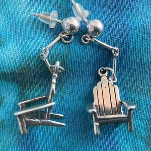 May include: Silver-toned Adirondack chair earrings. Each earring features a small chair charm suspended from a post with a silver ball and a connecting bar. The earrings are displayed against a blue and teal tie-dye background.