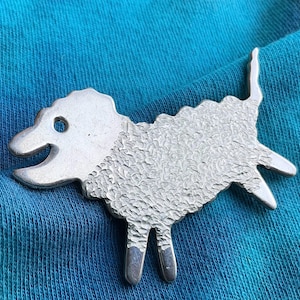 May include: A silver-colored sheep-shaped brooch with a textured woolly body and a smooth head, legs, and tail. The brooch is set against a blue fabric background. The sheep has a smiling expression and a hole for the eye.