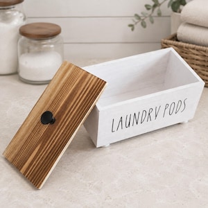 Può includere: Una scatola rettangolare in legno bianco con la scritta LAUNDRY PODS stampata in nero. La scatola presenta un coperchio rimovibile con finitura in venature del legno naturale e una piccola maniglia a pomello nera. Il contenitore poggia su quattro piedini.