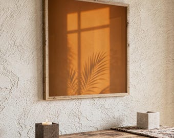 Pode incluir: Um espelho quadrado com moldura de madeira clara refletindo uma luz laranja quente de pôr do sol e a silhueta de folhas de palmeira. O espelho está posicionado sobre uma mesa de madeira rústica com um castiçal quadrado cinza escuro e um pote.