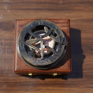 Puede incluir: Un reloj de sol circular de latón con brújula integrada, que presenta números romanos grabados y el texto Gilbert & Sons London. El instrumento está montado sobre una base cuadrada de madera con cierres de metal dorado en el borde frontal.