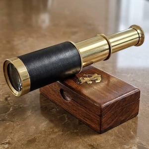 Peut inclure: Une longue-vue en laiton et cuir noir posée sur une boîte rectangulaire en bois. La longue-vue présente une finition en laiton poli avec une poignée en cuir noir. La boîte en bois a une finition brun foncé et une incrustation d'ancre en laiton.