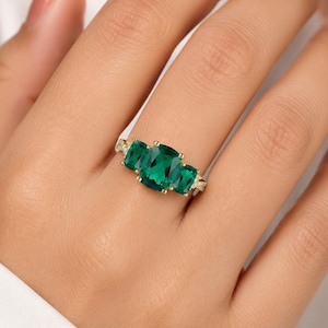 May include: A gold ring featuring a large cushion-cut green gemstone in the center, flanked by two smaller rectangular-cut green gemstones. The band is thin and accented with small, clear, sparkling stones along the sides of the setting.
