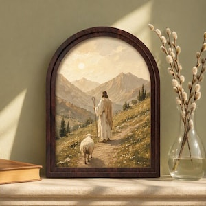 May include: An arched wall art print in a dark wood frame depicting a figure in a long robe holding a wooden staff, walking along a mountain path with a sheep. The scene features a sunlit landscape with rolling hills, distant peaks, and soft light.