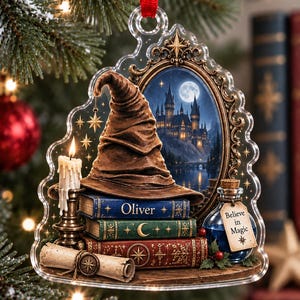 May include: A clear acrylic hanging ornament featuring a brown wizard hat resting on a stack of three books labeled Oliver, a lit candle, a scroll, and a blue potion bottle with a tag reading Believe in Magic, set against a castle backdrop.