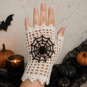May include: A fingerless glove made of cream-colored crochet yarn in a grid pattern, featuring a black crocheted spider web design with a small black spider at the center. The glove has a scalloped edge at the wrist and finger openings.
