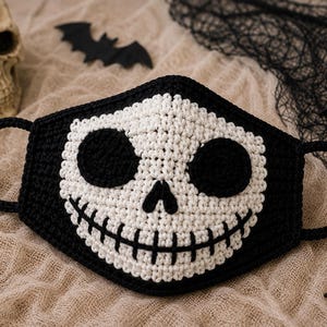 May include: A black and white crocheted face mask featuring a skull design. The mask has a white skull face with large circular black eye sockets, a small triangular nose, and a wide stitched mouth. It includes two black crocheted ear loops.