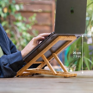 Può includere: Un supporto per laptop in bambù marrone chiaro con telaio angolato regolabile che sostiene un laptop grigio scuro. Il supporto presenta un design a doghe e un'altezza di 20 cm indicata da una linea tratteggiata bianca e testo a destra.