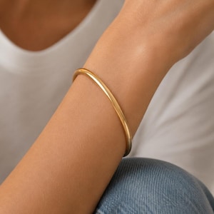 Op de afbeelding: Een gladde, gepolijste goudkleurige armband met een afgerond profiel. De armband heeft een doorlopend, minimalistisch cirkelvormig ontwerp dat comfortabel om de pols zit en een reflecterende metalen afwerking met een eenvoudige uitstraling.