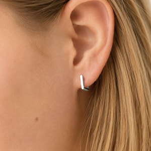 May include: A small, silver-toned hoop earring with a distinct rectangular or square-shaped profile. The earring features a polished, metallic finish and is secured through the earlobe, showcasing a clean, geometric design with sharp, defined edges.