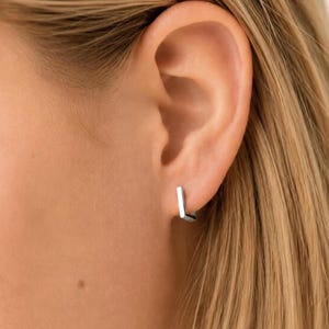 May include: A small, silver-toned hoop earring with a distinct angular, U-shaped design. The earring features a polished, metallic finish and is secured through the earlobe, showcasing a clean, geometric silhouette that sits close to the skin.