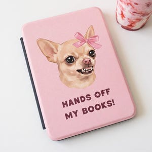 May include: A light pink e-reader cover featuring a graphic of a tan chihuahua wearing a pink bow on its head, baring its teeth, and the text "HANDS OFF MY BOOKS!" printed in dark brown letters below the dog illustration on the front cover.