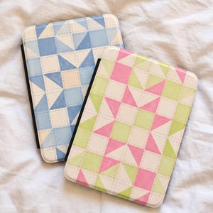 May include: Two rectangular e-reader cases with rounded corners. One case features a blue and white geometric quilt-like star pattern. The second case displays a pink, light green, and white geometric quilt-like star pattern with visible stitching.