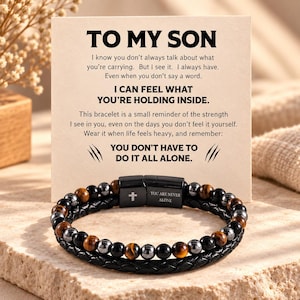 May include: A multi-strand bracelet featuring black braided leather and a row of alternating tiger eye and black beads. The black metal clasp displays a small cross and the engraved text YOU ARE NEVER ALONE on a card reading TO MY SON.