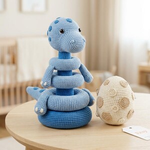 May include: A blue crocheted dinosaur stacking toy with a central post and three rings, featuring a rounded head with dark eyes and a tail. Beside it sits a cream-colored crocheted egg with tan circular patches and small metal accents.