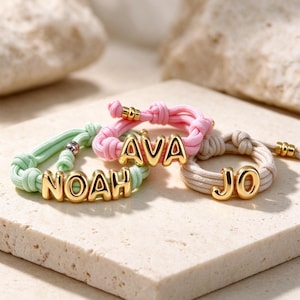 Op de afbeelding: Drie verstelbare koordarmbanden in lichtgroen, roze en beige. Elke armband is voorzien van goudkleurige metalen letterkralen die de namen NOAH, AVA en JO spellen. De armbanden zijn vastgezet met geknoopte koorden en kleine goudkleurige accenten.