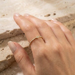 Peut inclure: Une fine bague en or ornée d'un petit motif floral à quatre pétales serti de minuscules pierres claires, complétée par d'autres pierres claires individuelles espacées le long de l'anneau en or poli et délicat, formant un bijou raffiné.
