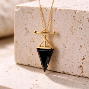 May include: A gold-tone necklace featuring a toggle clasp and a geometric pendant. The pendant consists of a triangular black stone set in a gold-tone frame with a pyramid-shaped top section, suspended from a delicate gold-tone chain.