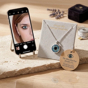 Puede incluir: Un collar con colgante de plata que presenta un diseño de iris azul realista sobre una tarjeta de presentación gris. La tarjeta dice Artisan Iris Portrait Pendants. Una etiqueta redonda dice The Watchful Eye Jewelry Co. Personalized Photo Pendant. Un teléfono inteligente muestra la foto de un ojo.