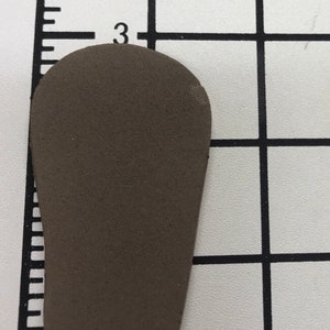 May include: A small, brown, teardrop-shaped object, approximately 2.5 inches long, rests on a grid background. The object has a smooth surface and rounded edges. The grid has markings for measurement.