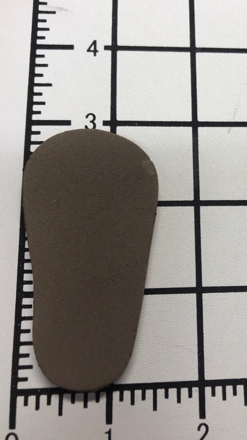 May include: A small, brown, teardrop-shaped object, approximately 2.5 inches long, rests on a grid background. The object has a smooth surface and rounded edges. The grid has markings for measurement.