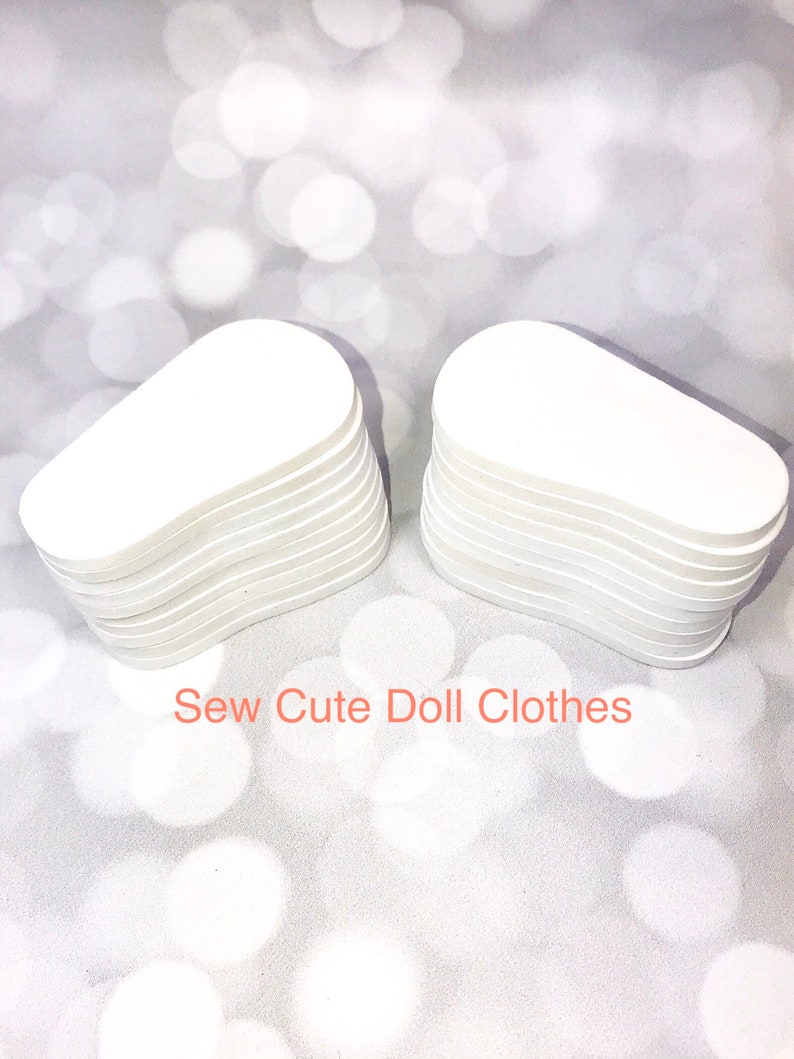 May include: Two stacks of white, oval-shaped doll clothes pieces. The pieces are stacked neatly, with visible edges. The text "Sew Cute Doll Clothes" is written in orange across the bottom of the image. The background is a soft, blurred, white and silver.