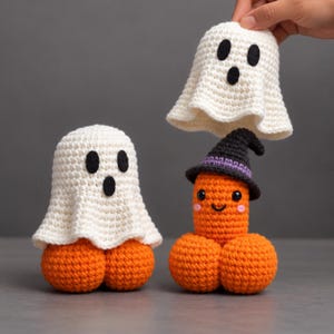 May include: Two crocheted figures featuring orange bases with round shapes. One figure is covered by a white ghost hood with black felt eyes and mouth. The other figure wears a black witch hat with a purple band, black eyes, and pink cheeks.