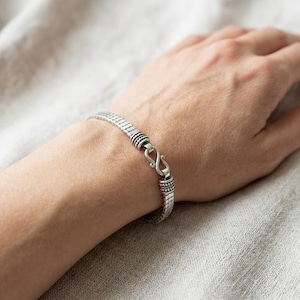 May include: A silver-toned bracelet featuring a flexible, segmented chain design with a textured finish. The bracelet is secured with a prominent S-shaped hook clasp, which connects to two decorative, ribbed cylindrical end caps on each side.