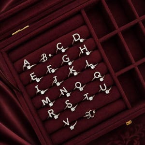 Può includere: Una collezione di anelli in metallo color argento, ognuno caratterizzato da una singola lettera dell'alfabeto dalla A alla Z. Ogni anello è impreziosito da piccole pietre chiare lungo la forma della lettera e include una pietra rotonda più grande.