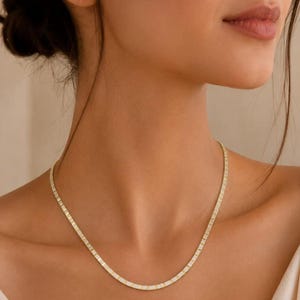 May include: A gold chain necklace featuring a series of rectangular links set with small, clear, sparkling stones. The necklace rests against the neck, showcasing a uniform design of metallic gold-toned metal and embedded white stones.