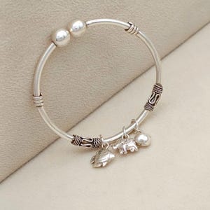 Puede incluir: Una pulsera rígida de tono plateado con dos cuentas esféricas lisas en la abertura y dos bandas metálicas decorativas texturizadas. Tres pequeños dijes de tono plateado cuelgan de la parte inferior, representando un pez, un elefante y una forma redondeada.
